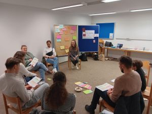 Zeigt eine Gruppe von 8 jungen Menschen zwischen 20 und 30 Jahren, die in einem Stuhlkreis sitzen. In der Mitte liegen Moderationsmaterialien aus, sie tauschen sich aus über Infos, die auf einem Blatt stehen, das sie in der Hand halten.