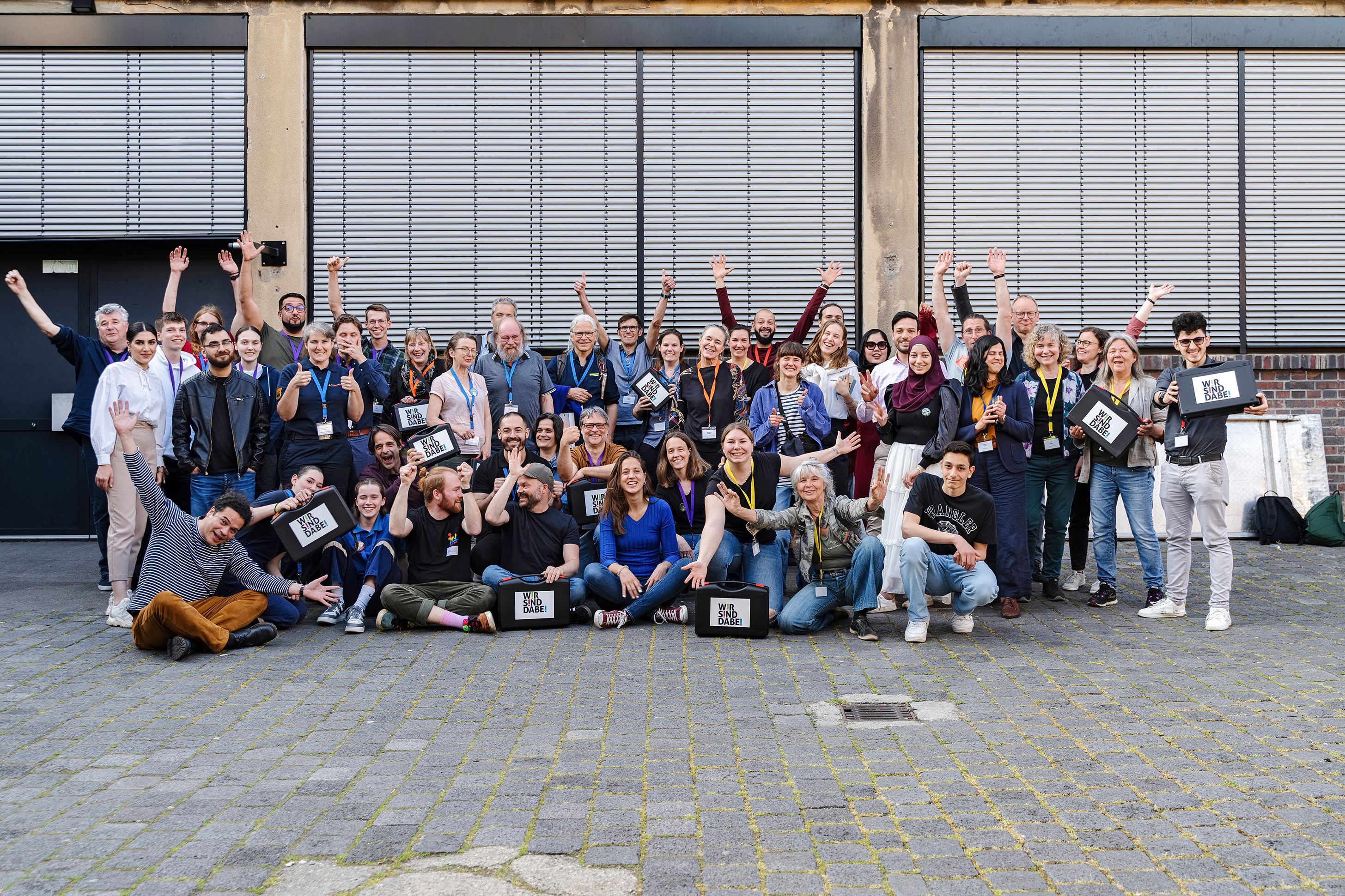 Ein Gruppenbild von vielen Menschen. Ein paar von ihnen halten Werkzeugkoffer mit Wir sind dabei-Aufdruck hoch.